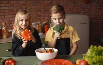 Cute girl and boy with fresh pepper and broccoli in hands standing near desk with assorted vegetables and salad bowl during vegetarian meal preparation