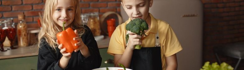 Cute girl and boy with fresh pepper and broccoli in hands standing near desk with assorted vegetables and salad bowl during vegetarian meal preparation