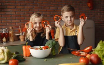 Two children holding capsicum slices while preparing vegetables in a cozy kitchen setting.