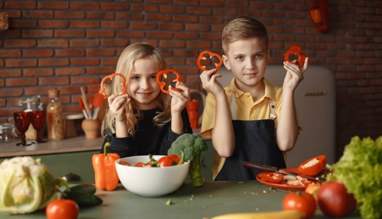 Two children holding capsicum slices while preparing vegetables in a cozy kitchen setting.